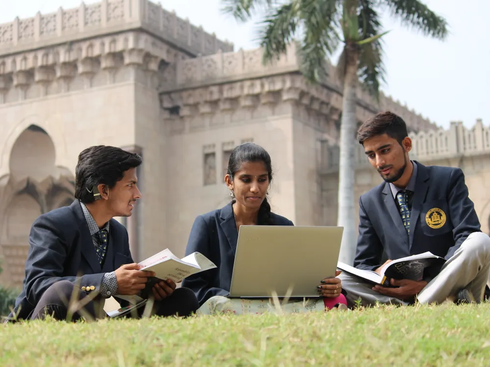 Three students working with a laptop and books on a college lawn